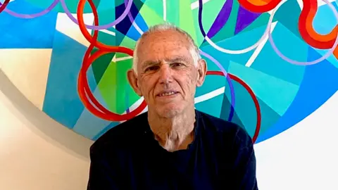 Tom Hume smiling into the camera, sitting in front of a circular geometric painting which is red, blue, white, green and purple. He has white, short hair and is wearing a dark long-sleeved top.