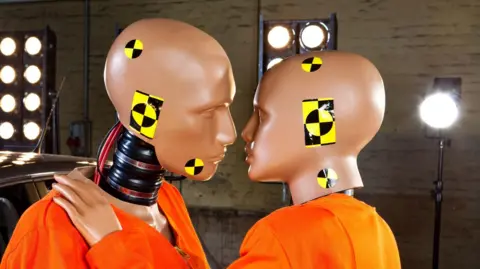 Two crash test dummies in an embrace with lights behind them 