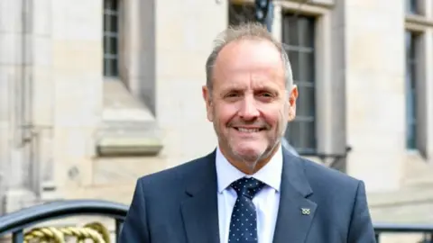 Hall is standing outside a cream-coloured building and is wearing a navy suit, shirt and tie. He has greying hair and a short beard.