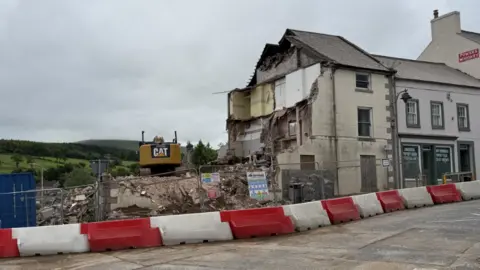 A large demolition site. Rubble is visible and an orange digger is sat atop a large pile of rubble