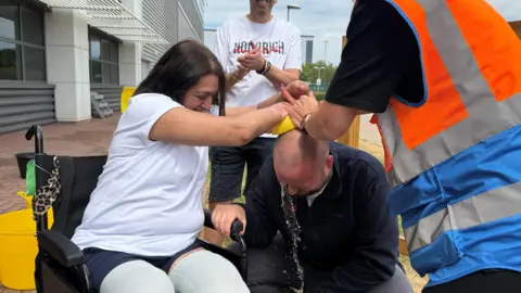 A woman wearing a white t-shirt is sitting in a wheelchair as she tips water over a bald man using a sponge. She is surrounded by people who are cheering her on. Beside her is a yellow bucket.