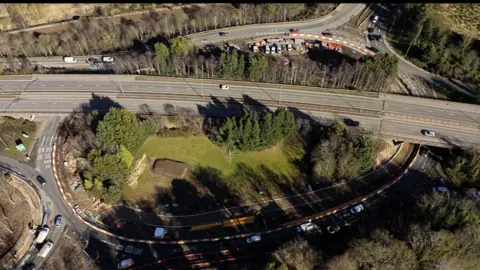 An aerial view of the Raigmore Interchange. It is a large structure featuring an oval shaped roundabout with a flyover going over the top of the roundabout and a grassy area.