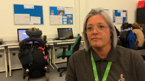 Hazel Wark is looking towards the camera, wearing a grey t-shirt and a green lanyard. She is in a classroom with a student in a wheelchair sitting behind her
