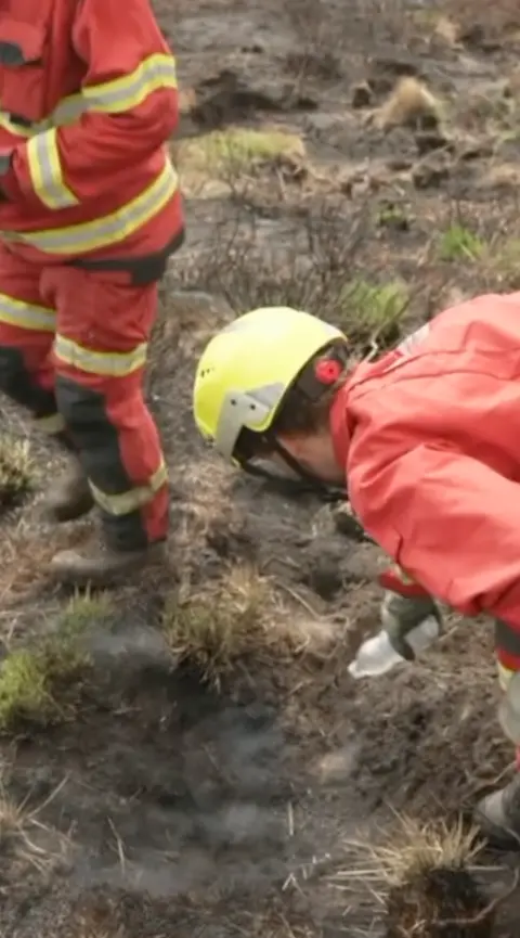 A man in protective wear pours water over a smoking peat on the moors.