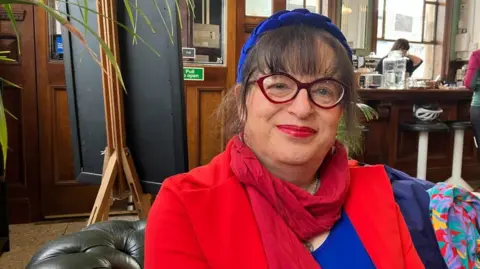 Vikki Irwin/BBC A woman is wearing a red jacket and scarf over a bright blue top. She is wearing a blue headband and dark red glasses. In the background, you can see the counter of a coffee shop.
