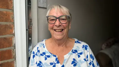 George King/BBC Jean McSorley wearing a blue and white floral top and glasses smiling into the camera while standing in her doorway.