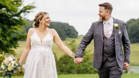 A newlywed couple hold hands and smile at each other against a backdrop of hills and trees. The groom is wearing a grey suit and the bride, who is holding some flowers in her other hand, is wearing a traditional white dress.