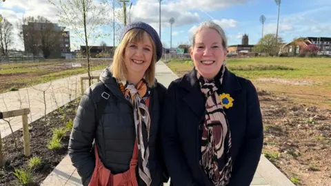 Two women stood next to each other, looking at the camera and smiling. They are wearing black coats and colourful scarves. Behind them is the pathway, with the cricket ground floodlights in the distance.