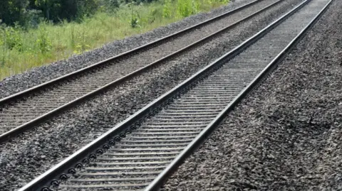 Two railway lines can be seen with gravel underneath and either side of them. Grassland is visible on the left side of the tracks.