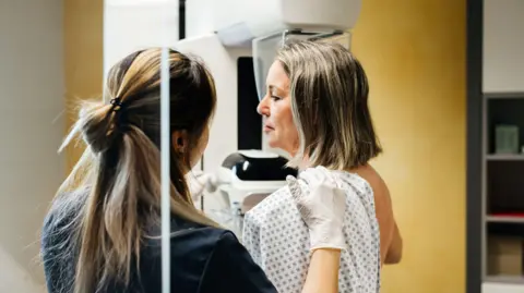 Healthcare professional wearing white examination gloves positioning a patient in a medical gown for a breast imaging scan using mammography equipment in a clinical setting.