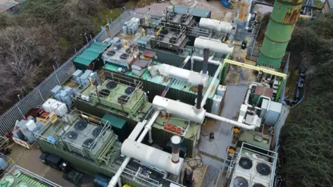 Melton CLP A drone shot of a number of large generator machines outdoors. There are large green containers with fans on the stop, connected by large white pipes. There is a silver metal fence around the perimeter, and a large green drum towering above the machines.