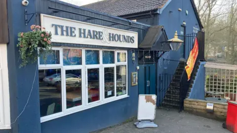 BBC A large blue pub with the Hare and the Hounds sign on the front. There is a hanging basket with pink flowers next to the sign and stairs leading up the outside of the building away from the camera. A white, blank advertising board stands outside.