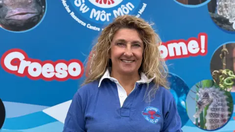 Anglesey Sea Zoo owner Frankie Hobro stands in front of a large blue sign for the zoo, with the word croeso on the left of the image. There is a logo above her head for the Sea Zoo, depicting a lobster graphic with Sea Zoo and Sw Môr Môn - with pictures of some of the aquatic animals either side. Frankie has long blonde wavy hair, and she is wearing a blue rugby style shirt with white collars