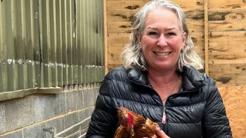 Katherine Ganczakowski/BBC Dawn French in a farm shed/barn, wearing a black puffa jacket and holding a brown hen. She has shoulder-length grey hair and is smiling broadly at the camera. Behind her can be seen a pile of hay.