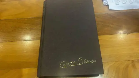 Westmeath Libraries A closed black book, in good condition, with the words Christy Brown in gold on the bottom right hand corner