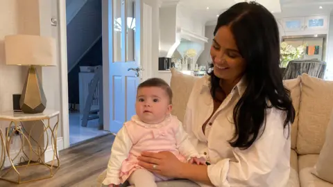 A dark-haired woman with a child on her knee. The woman is smiling and wearing a white shirt. The girl is looking forward and is wearing a pink pinafore, with a cream top and cream tights underneath. The couple is sitting on a brown sofa, with a gold table and lamp on one side and a light-coloured kitchen in the background.