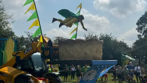 Man dressed in green and yellow superhero costume dives into a giant Yorkshire pudding, which sits on a JCB platform