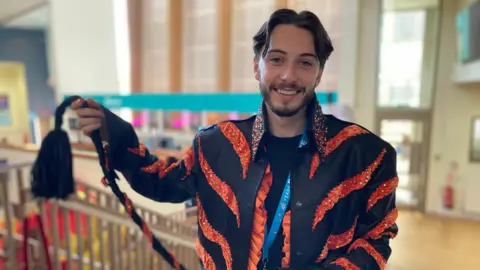 Alex Pope/BBC Oskar White, wearing a Sheer Kan, black and orange costume, holding its tail out. He is smiling and looking at the camera, and has short dark hair and a beard. A theatre's foyer is behind him. 