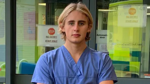 Finlay Maguire A young man in blue scrubs and blonde hair is standing in front of a hospital door. He is looking intently into the camera. 