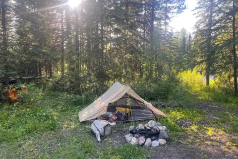 Naomi Hancocks A tent is pitched in woodland with stones around a fire in front of it