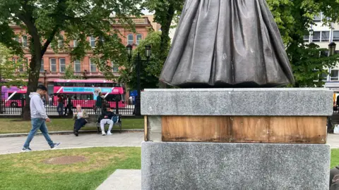 At the base of a statue, a section of plaque is missing, revealing a wooden panel. 
