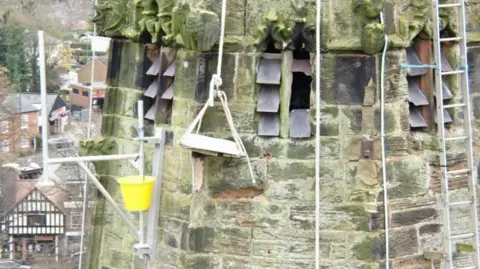 PA Media A wooden chair suspended by ropes with a yellow bucket also suspended nearby, both hanging from the side of a stone church steeple with small buildings visible below