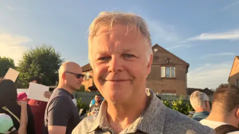 A head and shoulders image of Steve O'Dwyer smiling at the camera. He has short, grey hair and is wearing a grey polo shirt. He is standing outside in a crowd of people. There are houses in the background and the sky is blue.