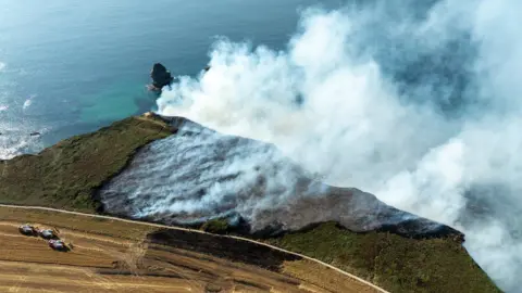 An aerial photo of Basset Cove, Portreath. There is a large amount of smoke on the cliff. The cliff is covered in grass. The sea is in the background. 