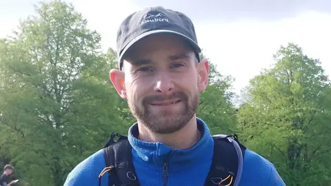 Michael wearing a black cap, blue half zip fleece and black running fence smiles at the camera. He has a dark beard and moustache.