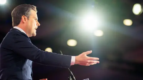 Getty Images Matt Gaetz delivers remarks at a podium, looking to the right of the picture. He has his right arm outstretched and wears a dark blue suit and white shirt. He appears to be on stage under strong spotlights.