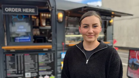 Charlie Rose/BBC Ally Nowell, wearing dark clothing with her hair tied back, stands in front of a coffee van and looks at the camera.