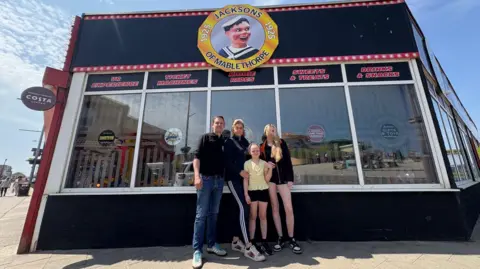 Gareth Cammack pictured with wife Kim and their two young daughters outside Jackson's Amusements in Mablethorpe. On the front of the building is the company logo containing a Laughing Sailor  