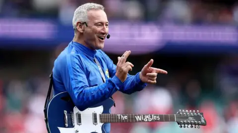 Matt King/Getty Images The blue wiggle on stage gesturing with his hands and holding a guitar.