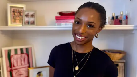 BBC/Hazel Shearing Tinuke Bamiro is wearing a black tshirt and gold jewellery and smiling into the camera. Behind her are shelves with perfumes, books, photos and prints on.
