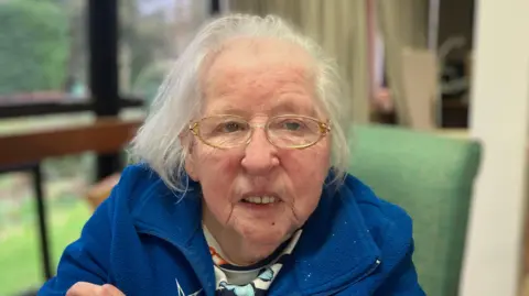 Sandra has short white hair and gold rimmed glasses.