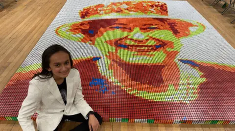 A boy sitting on the floor in front of a mosaic of Queen Elizabeth II made out of 3,000 Rubik's Cubes. He is wearing a white blazer and black trousers.