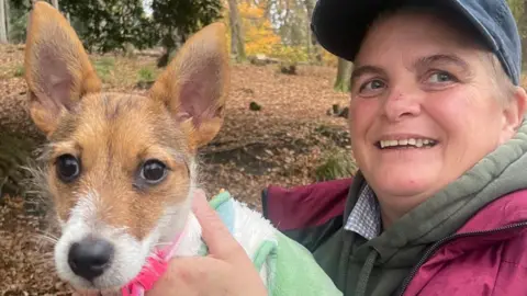 Ruth Parkinson A small brown and white dog is wearing a pink collar. It has pointy brown ears and is wearing a green jacket. It is being held by a woman wearing a blue cap and a pink jacket with a green hoodie. They are in a wooded area with brown leaves covering the floor.
