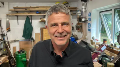 BBC/RICHARD EDWARDS A man standing in a shed and smiling at the camera. He has a moustache, fair hair and is wearing a black shirt. He is surrounded by tools.