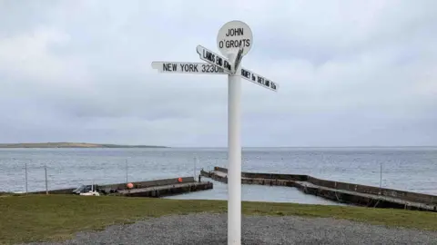 Barry Mackleston White sign reading 'John O Groats'. In the background is a large body of water.