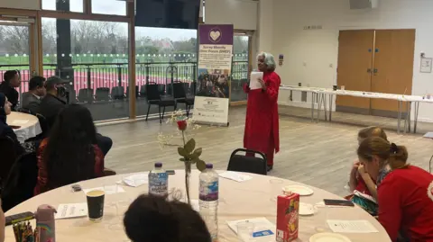 BBC/Julia Moore A woman dressed in red holding a microphone talking to attendees. 