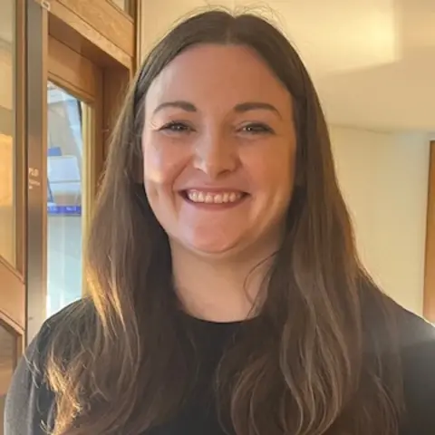 A smiling Natalie Don-Innes beside a bright window in the Scottish Parliament. She has long dark brown hair.