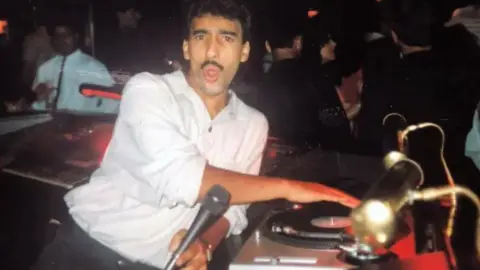 Moey Hassan A man, a DJ, looks into the camera as he spins a record. He is of South Asian heritage and has a moustache. There are people in the background. The photo is slightly faded.