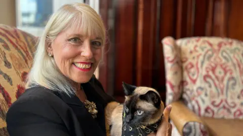 A woman, with blonde hair and wearing a black jacket, looking at the camera and smiling. She is sitting on a chair and holding a Siamese cat.