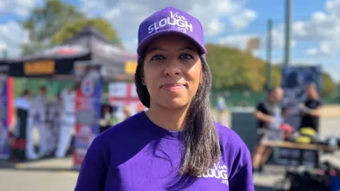 Amandeep is wearing a purple cap and t-shirt that said Viva Slough and is behind some stalls 
