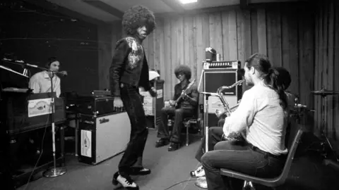 Getty Images Sly Stone sings and dances in the centre of a room with other members of the band pictured around him in a circle on various instruments. Two large amplifiers are dotted around the room.