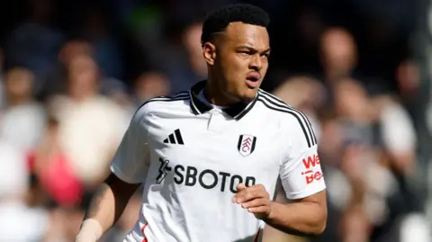 Getty Images Rodrigo Muniz playing for Fulham. It looks like he is running and leaning to his left. His mouth is open. He is wearing a white Fulham home kit with the black Adidas logo on the left with the three stripes across the shoulders. The fans in the stand are out of focus behind him.