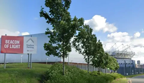 Google A red banner reading Beacon of Light, Stadium Parking, is perched on a grassland behind a row of young trees. A grey building is visible behind it with the Stadium of Light in the distance.