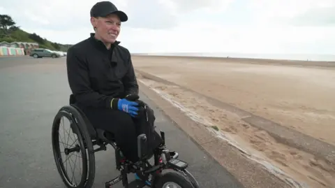 Wheelchair athlete Lexi Chambers sat at Exmouth seafront looking away from the camera smiling. She is wearing an all black outfit, with black glasses and a black cap. She is wearing blue gloves. 