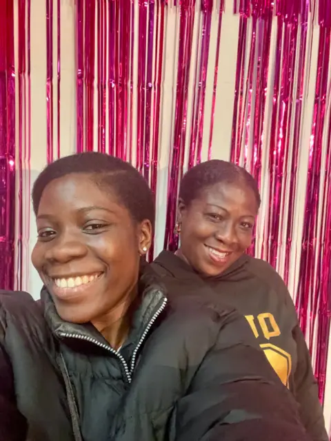 Mensah family Two women posing and smiling for a selfie. One is wearing a black jacket and the other is wearing a black hooded jumper with large yellow writing on the front.