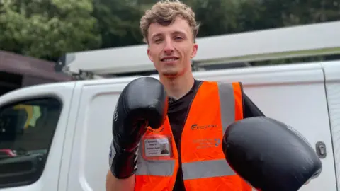 Simon Thake/BBC A young man stands next to a white van, wearing an orange jacket and black boxing gloves.